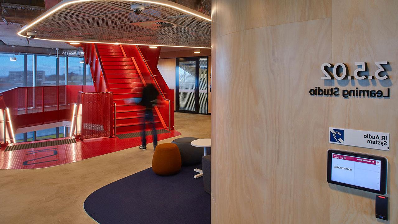 In the foreground is lettering on a wall that says "learning studio" with a student in the background walking up a staircase