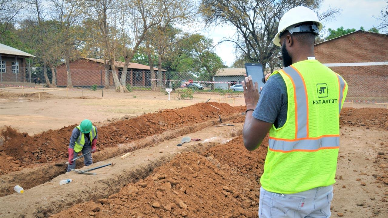 Man with a Tetra Tech high visibility vest holds a tablet and watches a man digging a trench in Malawi